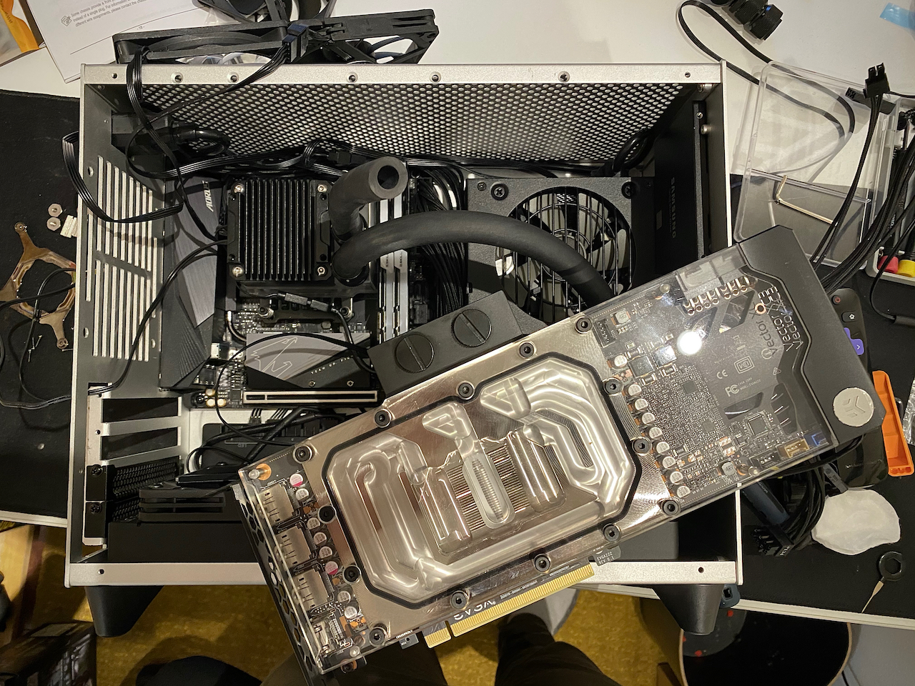 Graphics card with clear acrylic side visible showing the shiney nickel water block interior. Laying on top of the messy partially assembled computer.